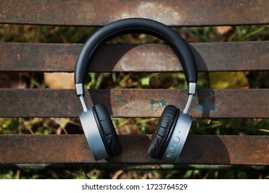 Set Of Black Leather And Metal Wireless Head Phones With Power And Volume Buttons On Earpiece Laying On Rusty Metal Bench In Autumn Outdoor Context