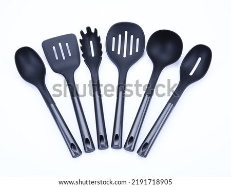 Set of black plastic cooking utensils consisting of kitchen skimmer, spatula, slotted spoon, kitchen spoon and soup ladle laid out on a white background