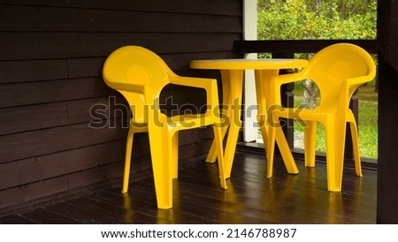 A set of a bright yellow plastic table and two chairs sits on a dark wood outdoor terrace in a nature house. High quality photo