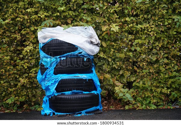 A set of\
car tires left or stored on the parking lot against a hedge. They\
are packed in a blue plastic bag decayed and torn. The tires are\
exposed. Micro plastic pollute\
environment.