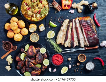 set of caribbean dishes, jerk pork belly, chicken curry, fried dumplings, roasted chicken thighs and drumsticks on plates on a black wooden table with spices and herbs, view from above, flat lay