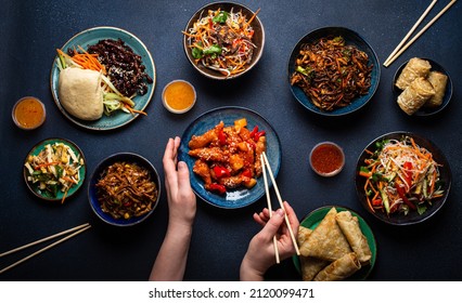 Set Of Chinese Dishes On Table, Female Hands Holding Chopsticks: Sweet And Sour Chicken, Fried Spring Rolls, Noodles, Rice, Steamed Buns With Bbq Glazed Pork, Asian Style Banquet Or Buffet, Top View 
