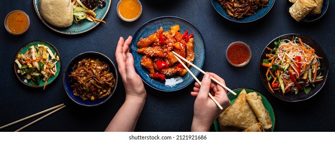 Set of Chinese dishes on table, female hands holding chopsticks: sweet and sour chicken, fried spring rolls, noodles, rice, steamed buns with bbq glazed pork, Asian style banquet or buffet, top view 