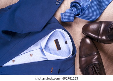 set of classic mens clothes - blue suit, shirts, brown shoes, belt and tie on wooden background. Men's accessories set. Top view. Copy space for text.