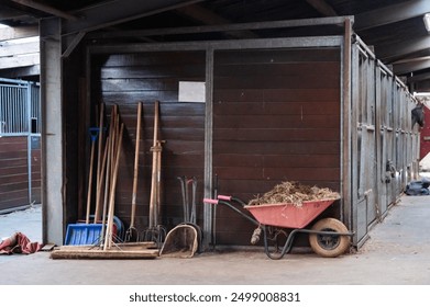 A set of cleaning tools and a red wheelbarrow by a stable aisle - Powered by Shutterstock