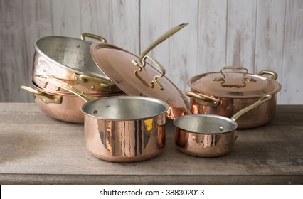 A set of copper cookware that include a small and medium-sized saucepan, a large saucepan, pot, and cover stacked on top of the other, and a covered small pot, all placed on a wooden countertop.