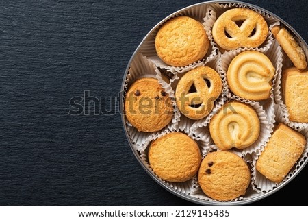 Set of crispy shortbread biscuits in an open tin over black slate surface. Tasty danish butter cookies in can for breakfast. Baked pastry, sweet food and calories concepts. Copy space. Top view.