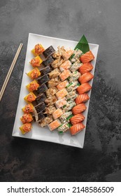 Set Of Different Colorful Maki Sushi Rolls With Tuna, Salmon, Shrimp, Crab And Avocado And Green Bamboo Leaf In A White Ceramic Plate With Chopstick On A Dark Gray Textured Background, Top View