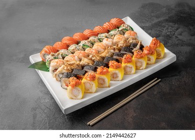 Set Of Different Colorful Maki Sushi Rolls With Tuna, Salmon, Shrimp, Crab And Avocado And Green Bamboo Leaf In A White Ceramic Plate With Chopstick On A Dark Gray Textured Background, Side View