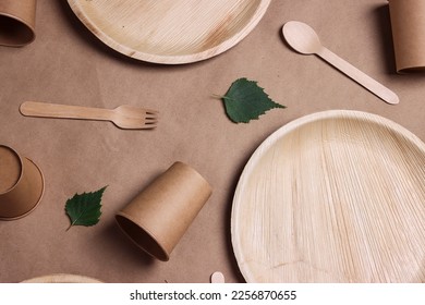 Set of different eco-friendly tableware with green birch leaves on brown paper  background. Bamboo plates, paper cups and wooden cutlery. Zero waste, plastic free, disposable tableware. Top view.