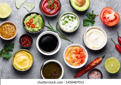 Set Of Different Sauces In Bowls And Ingredients On Gray Rustic Concrete Background, Top View. Tomato Ketchup, Mayonnaise, Guacamole, Mustard, Soy Sauce, Pesto, Cheese Sauce - Assortment Of Dips 