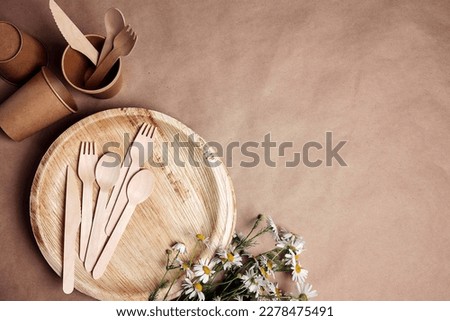 Set of eco-friendly tableware with bouquet of daisies on brown paper background. Bamboo plates, paper cups and wooden cutlery. Zero waste, plastic free, biodegradable, disposable tableware. Top view.