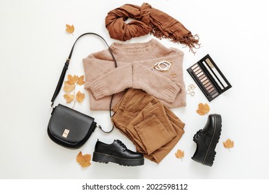 Set Female Autumn Clothing On White Background. Stylish Woman Outfit In Trendy Brown Earthy Color With Bag Cosmetics Accessories And Autumn Leaves.