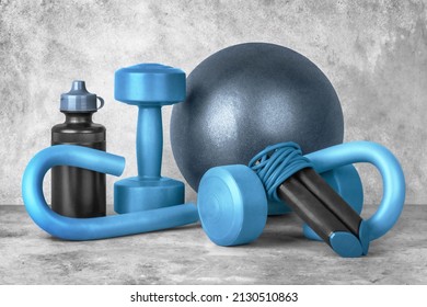 Set of fitness equipment on a gray concrete background, front view, close-up. Dumbbells, sport bottle of water, jump rope, s-shaped leg exercise machine, fitness ball. Home workout.