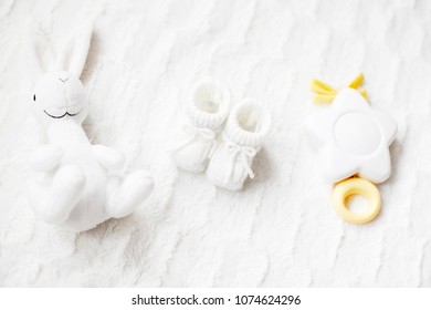 A Set Of Folded White Baby Clothes With Shoes And Toys On A White Blanket