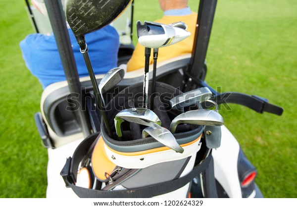 Set of golf clubs in sports bag on back\
of golf car being driven by one of mature\
players