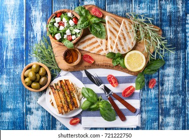 Set Of Greek Mezze: Grilled Halloumi Cheese, Pita Bread, Grilled Bell Peppers, Green Olives And Feta With Greens Decorated With Herbs, Lemon And Olive Oil Over A Blue Wooden Board. Top View