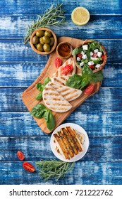 Set Of Greek Mezze: Grilled Halloumi Cheese, Pita Bread, Grilled Bell Peppers, Green Olives And Feta With Greens Decorated With Herbs, Lemon And Olive Oil Over A Blue Wooden Board. Top View