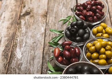 A set of green, red and black olives on  background. Various types of olives in bowls and fresh olive leaves. Vegan. Olive fruits. Place for text. Copy space. - Shutterstock ID 2380444781