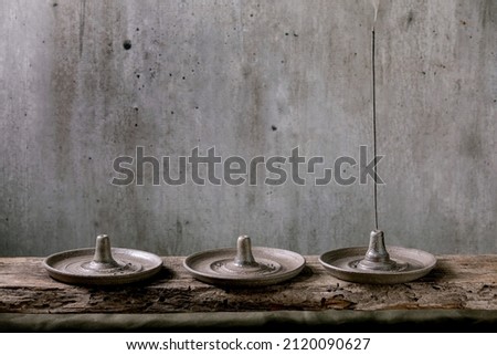 Set of hand crafted gray ceramic incense holder with fuming incense stick on wooden table with grey wall at background.