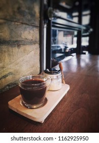 Set Of Hot Coffee (Caffe Americano) With Coffee Creamer In Glass Jar On Wooden Table Counter Bar With Grunge And Rough Concrete Wall Background In Cafe And Restaurant