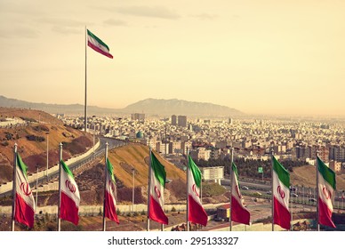 Set of Iran flags in Front of Tehran Skyline and one large flag in the background at sunset with orange warm tone.