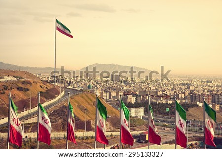 Set of Iran flags in Front of Tehran Skyline and one large flag in the background at sunset with orange warm tone.