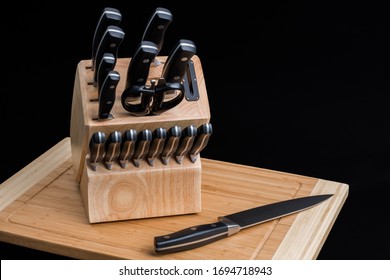 Set Of Kitchen Knives In A Knife Block With The Chef Knife Laying On A Cutting Board, Isolated On A Black Background