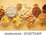 Set of many bowl with different salty snacks on bright yellow background. Chips, popcorn, nuts, nachos and pretzels. Party food. Top view