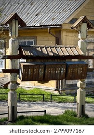 A set of old, weathered mailboxes from the Soviet era is seen in Lithuania. The mailboxes are rusted and have multiple compartments, each numbered. They are mounted on a wooden frame with a corrugated metal roof, positioned in front of a wooden house with peeling paint and a corrugated roof. The surroundings include grass and a paved walkway. These structures are characteristic of Soviet-influenced architecture commonly found in Eastern Europe. A rustic wooden structure supports a row of metal mailboxes, sheltered by a small, corrugated metal roof. The mailboxes are numbered and appear well-used, indicating service to multiple households. In the background is a weathered wooden building with a corrugated roof, typical of rural architecture. The scene is set in a grassy area with a pathway and simple metal fencing. This setup suggests a communal living or multi-family property in a rural setting.