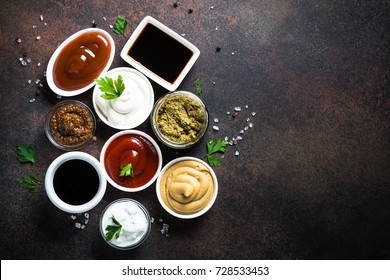 Set Of Sauces - Ketchup, Mayonnaise, Mustard Soy Sauce, Bbq Sauce, Pesto, Mustard Grains And Pomegranate Sauce On Dark Rusty Stone Or Metal Background. Top View Copy Space.