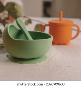 Set Of Silicone Baby Dishes. Green Bowl, Spoon And Orange Sippy Cup On Table Against Background Of Eucalyptus Sprig. Creative Blur And Grain. Square Frame Instagram Use