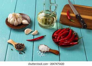 Set Of Spices On A Rustic Turquoise Wooden Table. Red Hot Chili Peppers, Garlic On A Wooden Saucer, Sea Salt And Black Peppercorns In Wooden Spoons And Olive Oil Glass Jar. Front View.