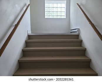 A set of stairs leading up to a landing. There is a large window on the landing and a small, metal heating unit along the wall. There are wooden banisters and rubber anti-slip covering on the steps.