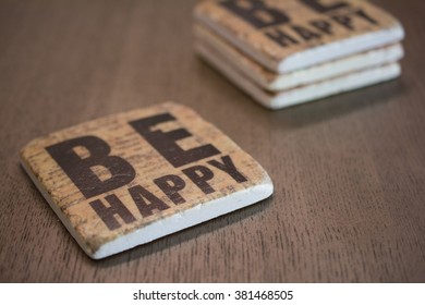 A set of stone coasters stacked on a wooden table surface reading Be Happy.