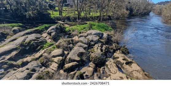 Set of stones in a river, trees, grass and bushes on their sides, houses in the background and blue sky