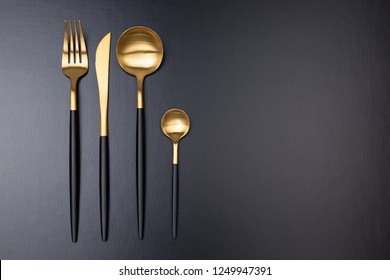 Set of stylish black and gold cutlery on black background. Dark and moody vibes. Fashionable and luxury eating. Flat-lay, top view. Copy space for your text.