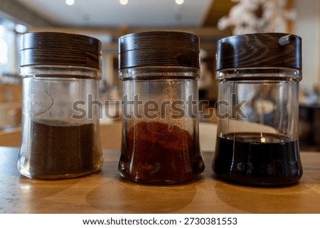 Set of three spice containers on wooden table: ground black pepper, red chili powder, and soy sauce in glass jars with wooden lids