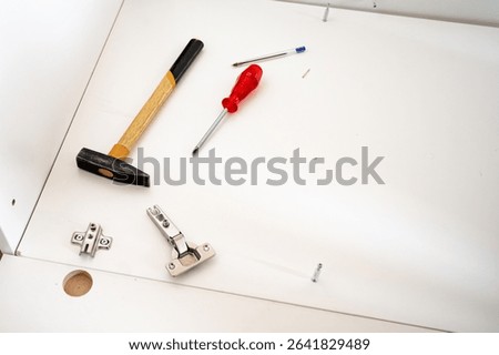 Set of tools and hardware for cabinet assembly, including a hinge, hammer, screwdriver, and screws on a white surface