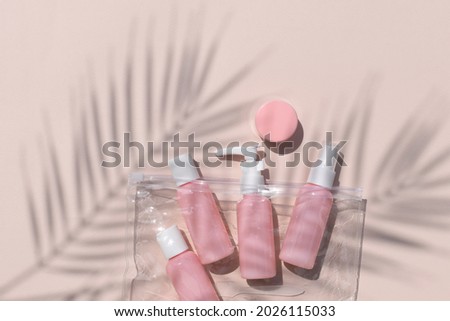 Set of travel size pink cosmetic bottles out clear bag. Mock up bodycare products for vacation or journey. Palm leaves shadows on sunlit neutral background. Feminine flat lay with copyspace.
