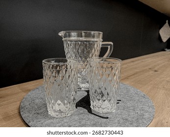 A set of two clear diamond-patterned glasses and a matching pitcher on a gray felt coaster, creating a simple yet elegant aesthetic for a kitchen or dining table, with a wooden background.