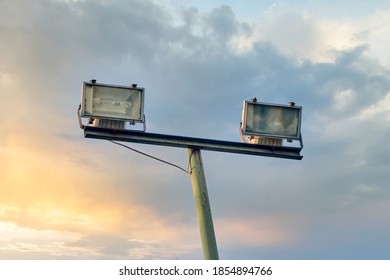 Set Of Two Outdoor Floodlight And A Pole For Sport Stadium, Car Parking Lot Or Public Park Illumination Against Beautiful Sunset Sky And Clouds Background With Copy Space.