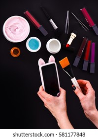 Set Of Various Cosmetics: Cream, Gel, Lipsticks, Lip Gloss, Brush, Powder Puff, Tweezers And Cell Phone In Hands Isolated On Black Background, Copy Space, Top View