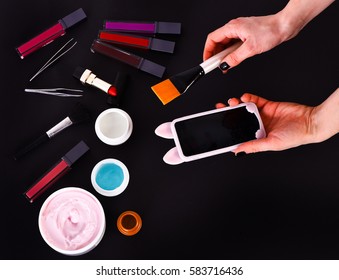 Set Of Various Cosmetics: Cream, Gel, Lipsticks, Lip Gloss, Brush, Powder Puff, Tweezers And Cell Phone In Hands Isolated On Black Background, Copy Space, Top View