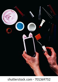 Set Of Various Cosmetics: Cream, Gel, Lipsticks, Lip Gloss, Brush, Powder Puff, Tweezers And Cell Phone In Hands Isolated On Black Background, Copy Space, Top View