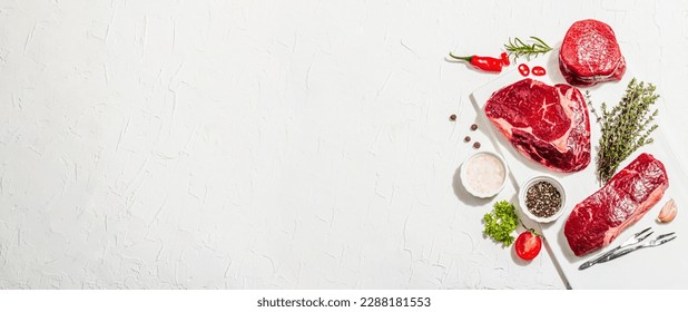Set of various steaks with spices and herbs. Classic raw meat cuts includes ribeye, eye round and striploin steaks. Plaster white background, flat lay, banner format - Powered by Shutterstock