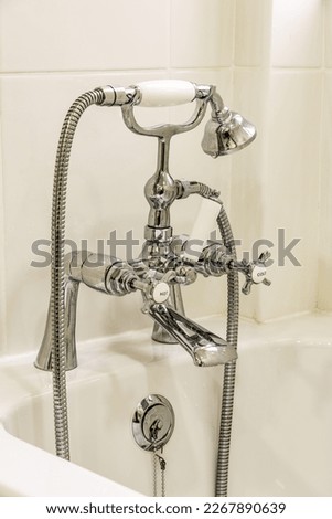 Set of Victorian style chrome finish, deck mounted, bath taps with shower resting in a cradle with tiled wall behind