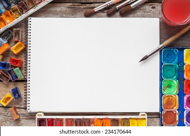 Set of watercolor paints,  brushes for painting and blank white paper sheet of sketchbook on vintage wooden background. Top view. - Powered by Shutterstock