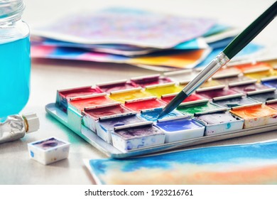 Set of watercolor paints, paintbrush, glass of water and paper sheets of watercolor paintings on background. Selective focus. 