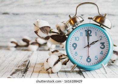 Set Your Clocks Forward And Spring Ahead With This Clock And Bolt Of Cotton Over A White Wooden Table. Daylight Saving Time Concept. Selective Focus With Blurred Background.